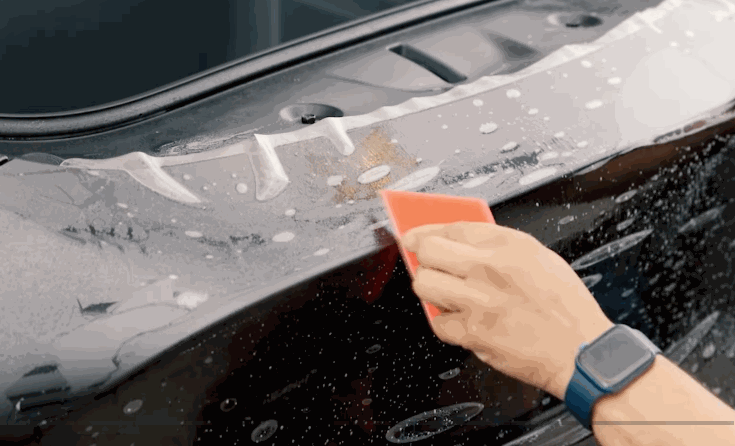 A technician uses a small squeegee to apply paint protection film around the headlight of a black vehicle. Water droplets cover the panel as the clear film is shaped into place.