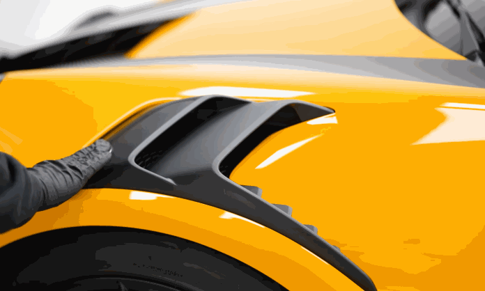 A technician in black gloves cleans the side vent on a yellow sports car during car detailing. The close up shows careful work around the black exterior trim.