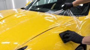 Technician applies paint protection film to the hood of a yellow car using a squeegee.