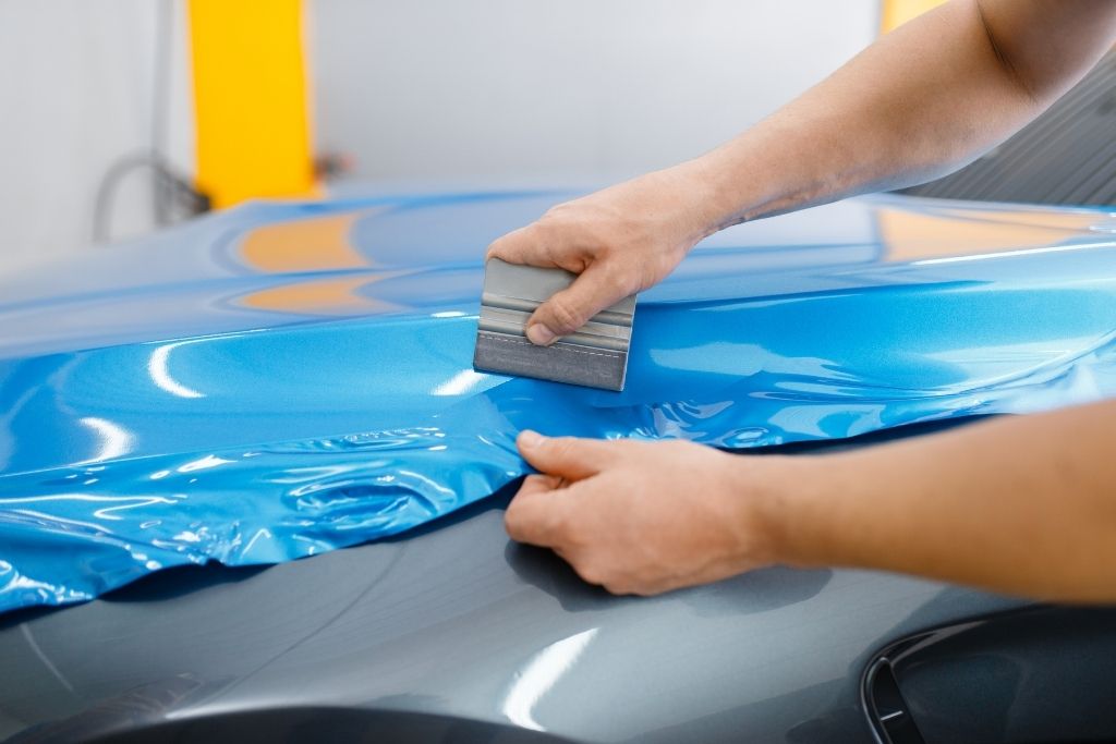 Technician applying a glossy blue vinyl wrap to a vehicle hood using a squeegee during professional car wrap installation.