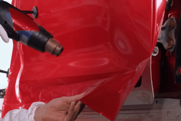 Technician heating and stretching glossy red vinyl wrap over a vehicle body panel during installation.