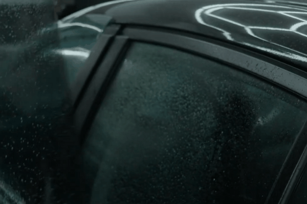 Close-up of a car door and rear window with freshly applied window tint film, showing moisture droplets and reflective light patterns from indoor lighting.