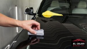 Hand applying ceramic coating to a glossy black car hood with an applicator pad while a yellow microfiber towel rests nearby. The reflective finish highlights the smooth protected surface.