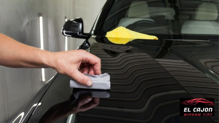 Hand applying ceramic coating to a glossy black car hood with an applicator pad while a yellow microfiber towel rests nearby. The reflective finish highlights the smooth protected surface.