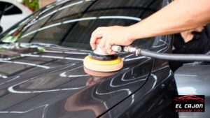 A technician using a machine polisher on a dark colored car panel to refine the surface during paint correction. The spinning pad works across the paint to remove swirl marks and enhance shine while a logo reading "EL CAJON WINDOW TINT" appears in the corner.