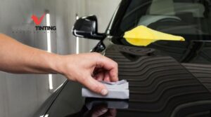 Hand applying ceramic coating to a glossy black car hood with an applicator pad while a yellow microfiber towel rests nearby. The reflective finish highlights the smooth protected surface.