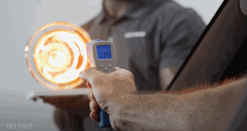 Technician uses a handheld device to measure heat from a bright lamp labeled "NO TINT" in the corner, demonstrating how window tint reduces heat and glare. The digital screen displays a temperature reading of "114".