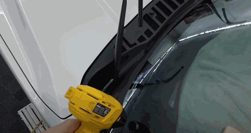 Close up of a heat gun being used on window tint along a car windshield, helping the film shrink and conform smoothly to the curved glass surface.