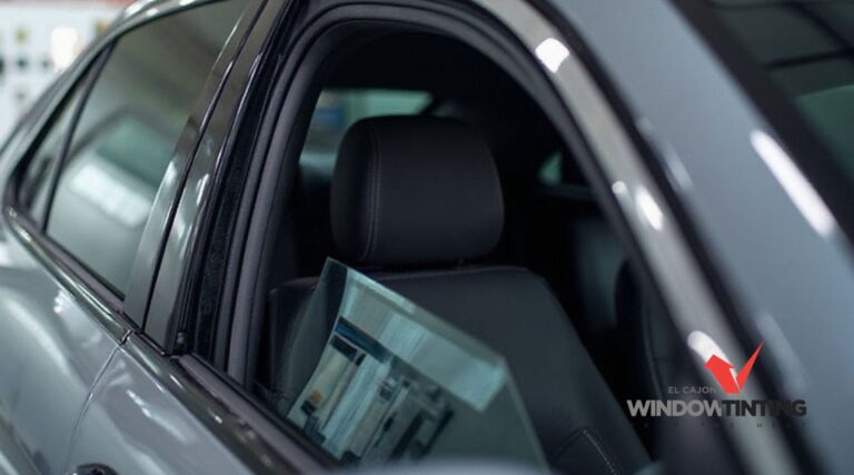 Gray sedan with newly installed window tint on the side windows parked inside a shop, with the logo "EL CAJON WINDOW TINT" visible in the top left corner. The slightly lowered window shows the clean edge and professional finish of the tint.