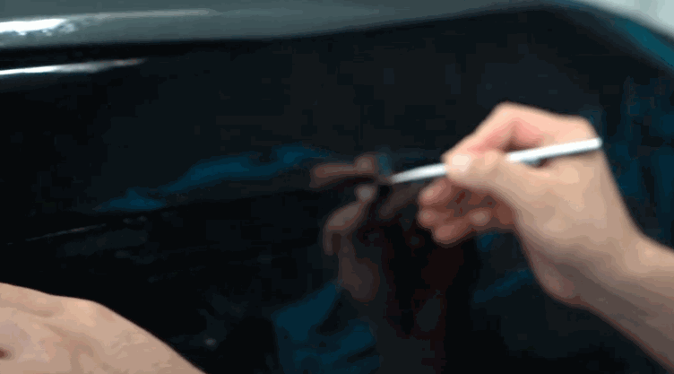 A technician marks dark window tint on a car window with a white pencil before trimming. The glass reflects the installer’s hand and tool.