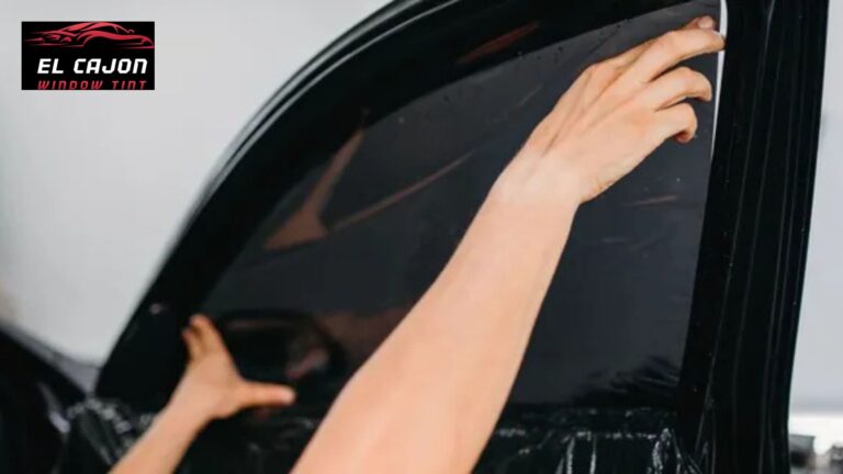 Hands smooth dark window tint onto a car side window near the upper corner. The logo in the corner reads "EL CAJON WINDOW TINT".