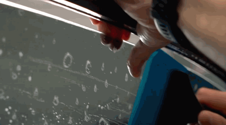 A technician trims window tint along the top edge of a wet car window with a red cutting tool. Small water bubbles cover the glass during installation.