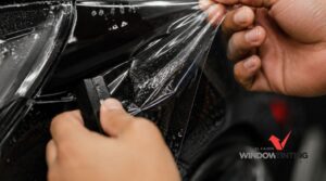 A technician applies paint protection film around the headlight and front panel of a black vehicle. The text “EL CAJON WINDOW TINT” appears in the top left corner.