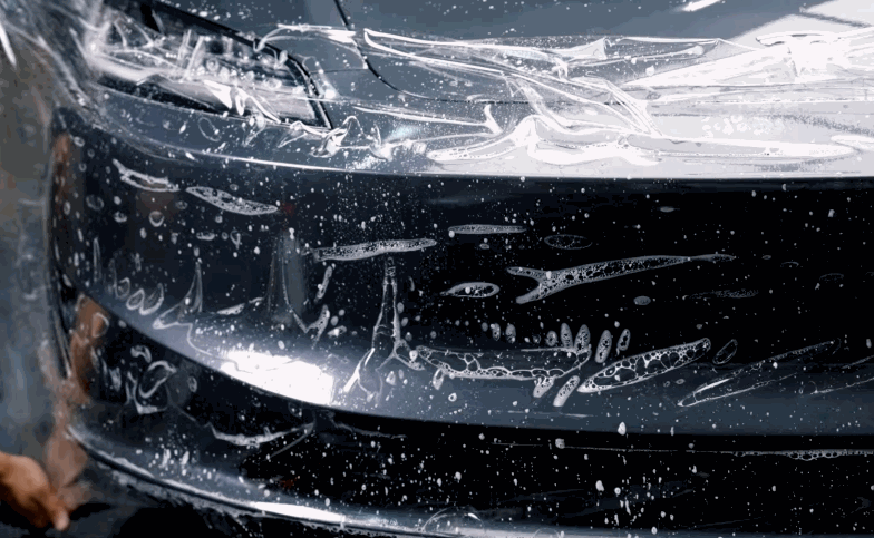 Close-up of paint protection film being laid over the front bumper of a black vehicle.